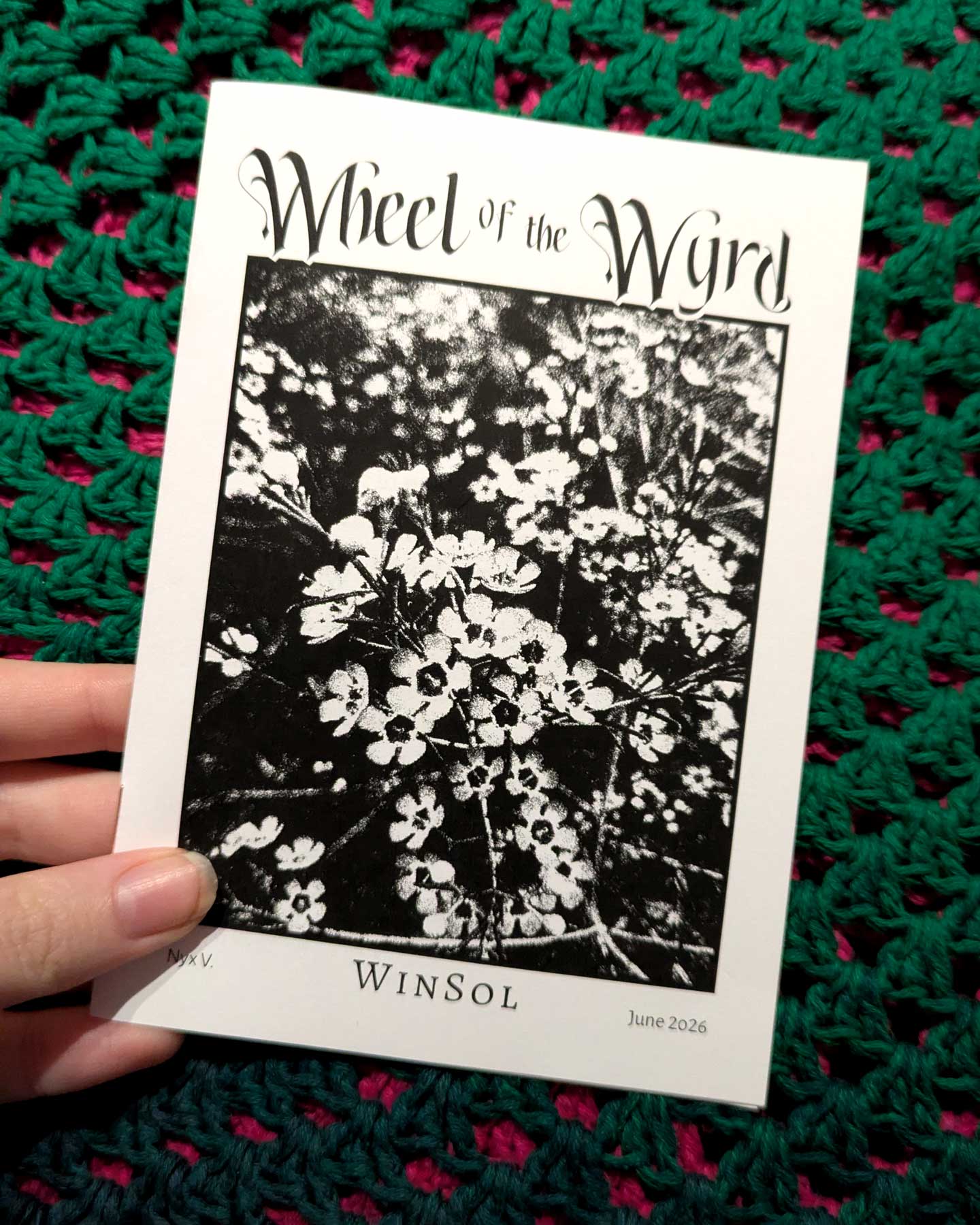 A hand holding a printed copy of the WinSol zine in front of a crocheted background. 