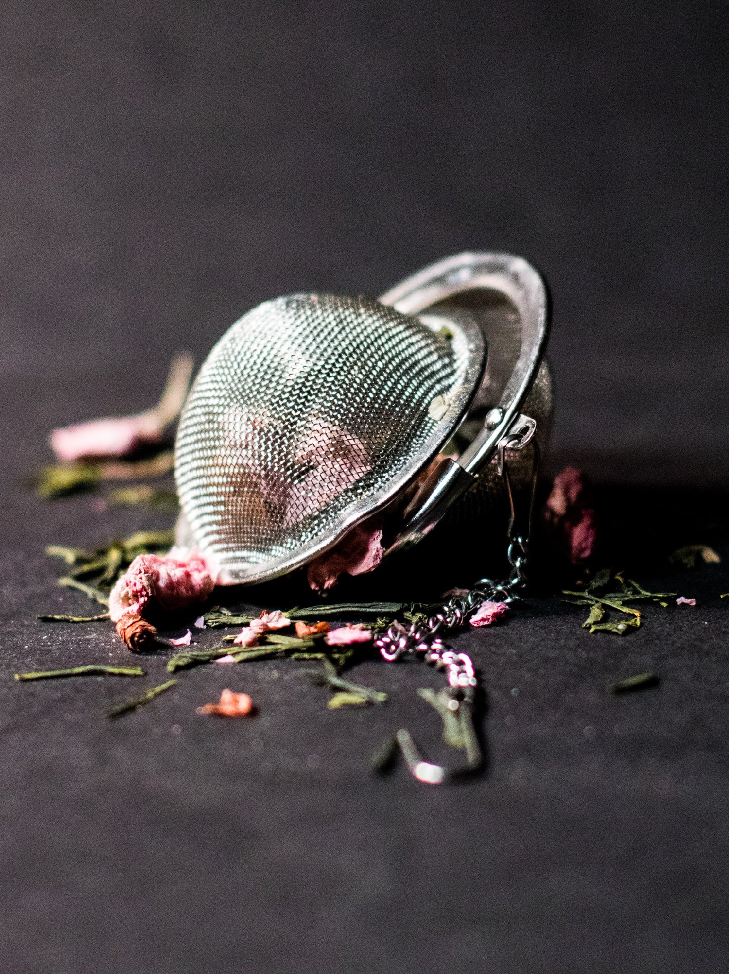 A mesh tea ball, slightly open, with green tea leaves and rose petals spilling from it