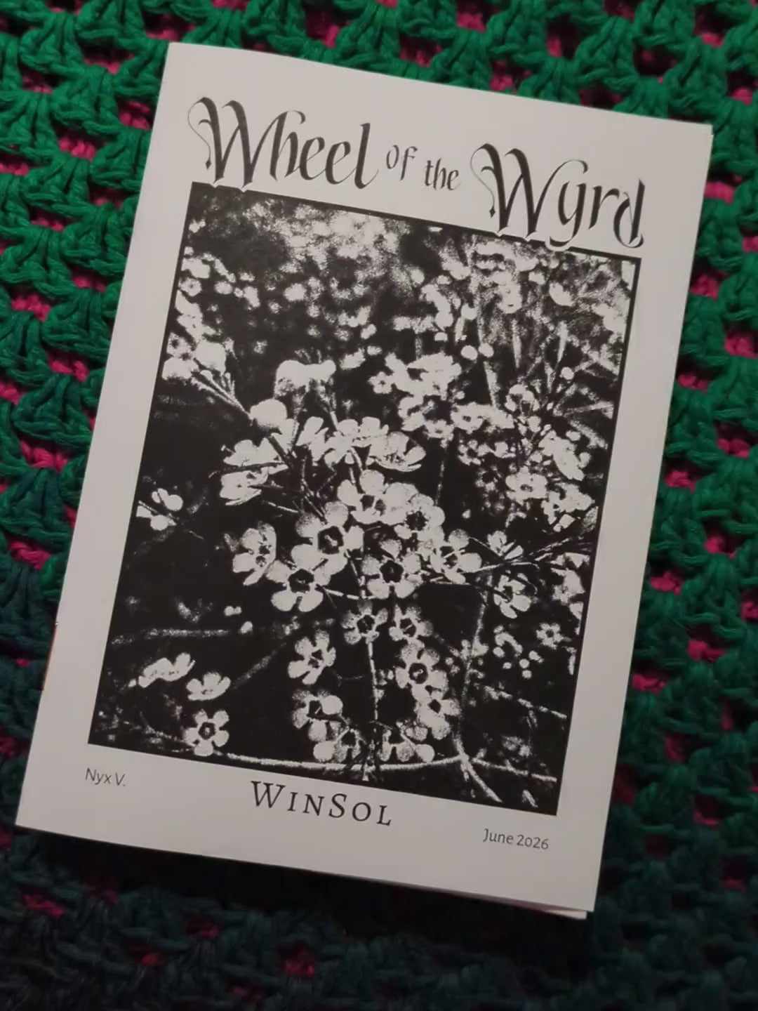 A hand flipping through a printed copy of the WinSol zine, in front of a crocheted background. 