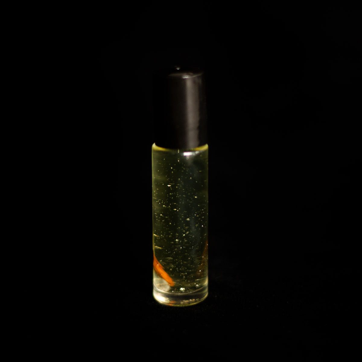 A perfume vial filled with pale yellow oil, containing a sandalwood chip, a small clear quartz crystal chip, and bio-glitter