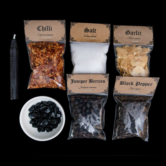 A black chime candle, small bowl of black tourmaline crystal chips, and 5 bags of herbs with Victorian-apothecary-style brown labels at the top give the common and Latin names